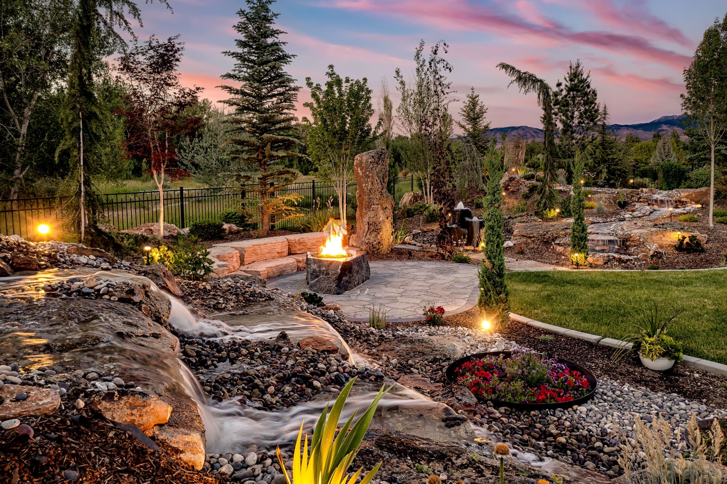 Residential landscape at dusk with a cascading water feature and stone fire bowl